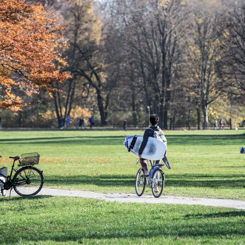 Person fährt mit dem Fahrrad durch einen Park und hat ein Surfboard unter dem Arm